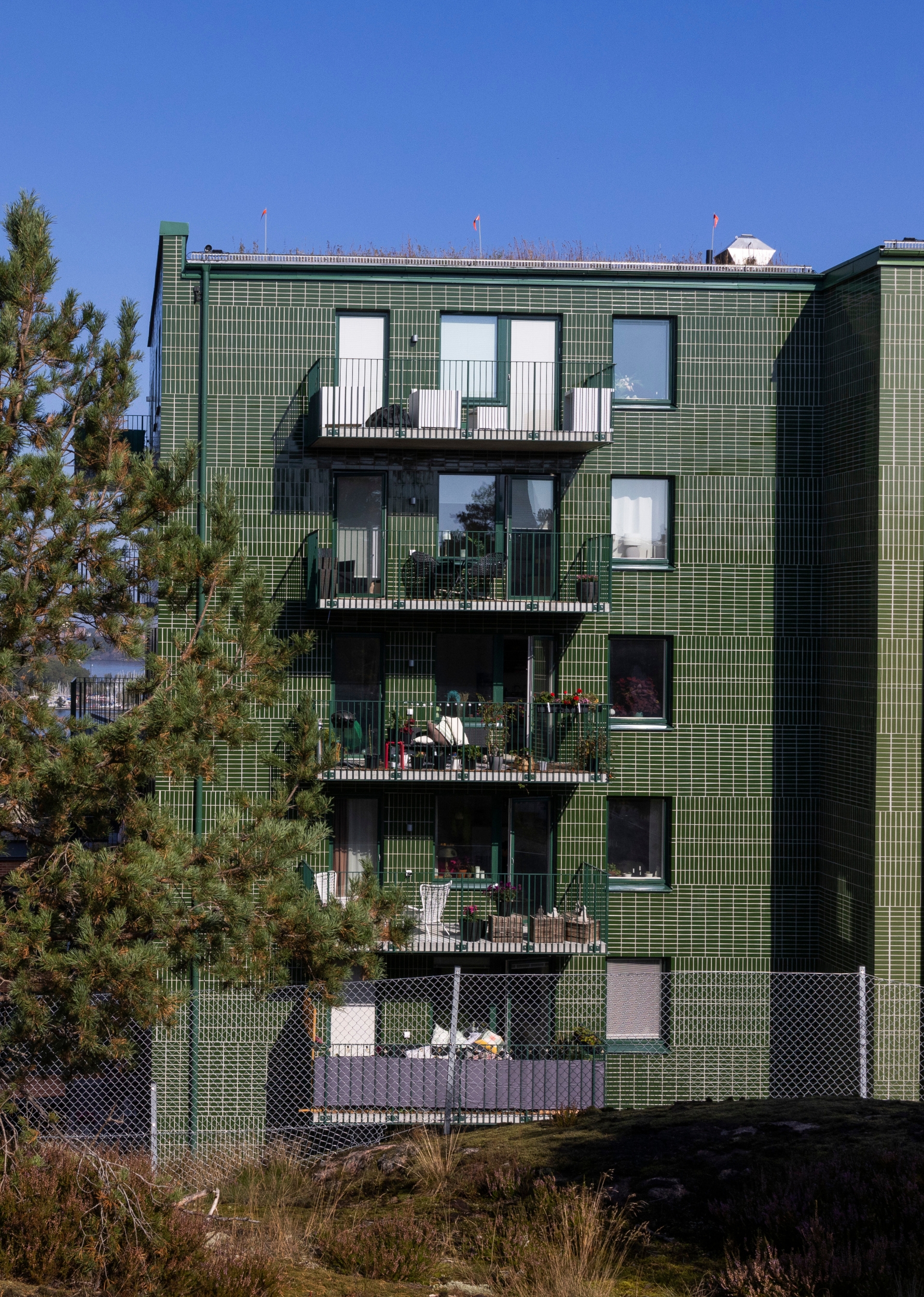 Flerbostadshus med fasad klädd i grönt kakel i ett grafiskt rutmönster. Flera balkonger med möbler och växter, mot en blå himmel.