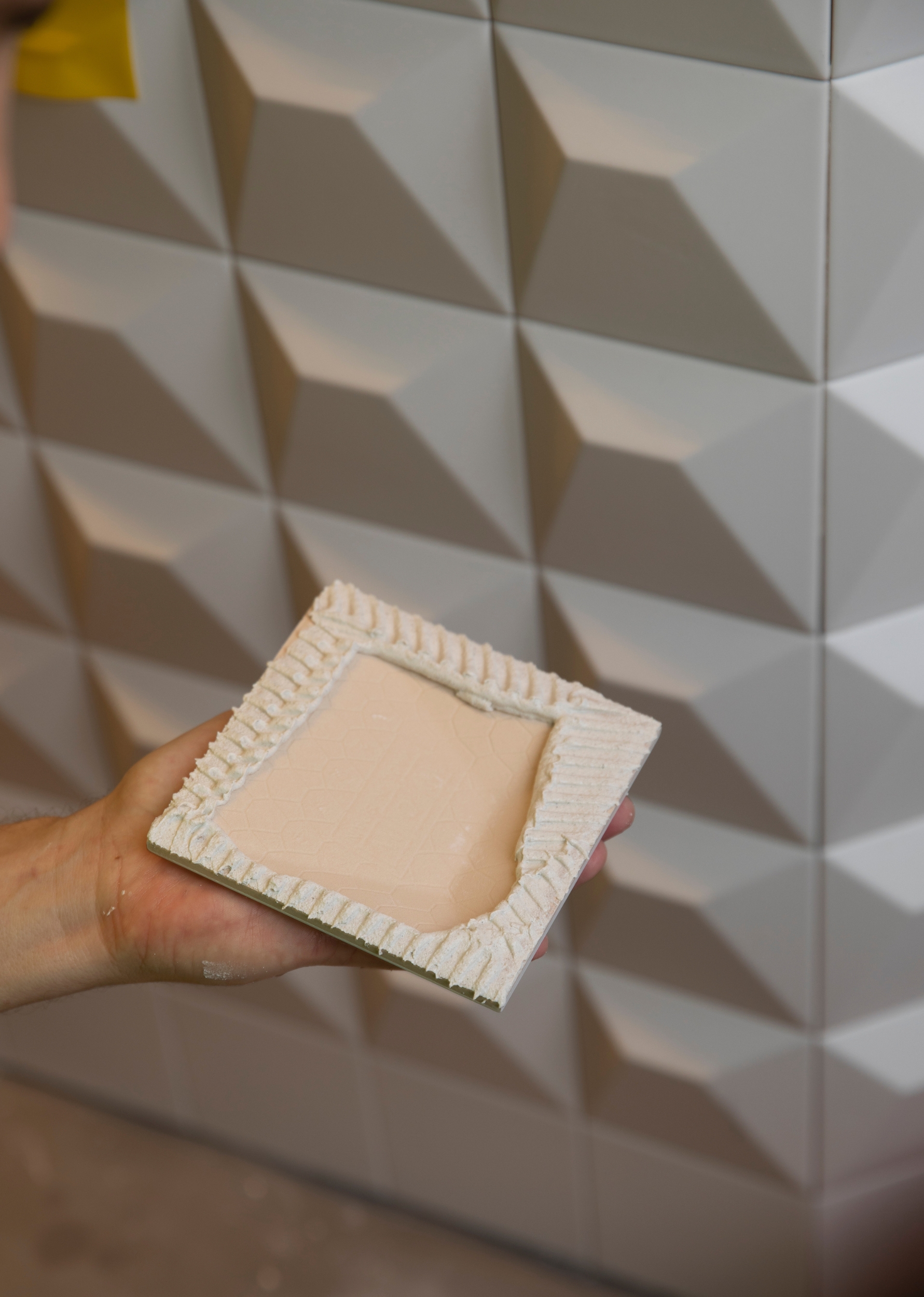 En hand håller upp en oglaserad dekorplatta med reliefmönster, framför en vägg klädd i vita, geometriskt formade kakelplattor.